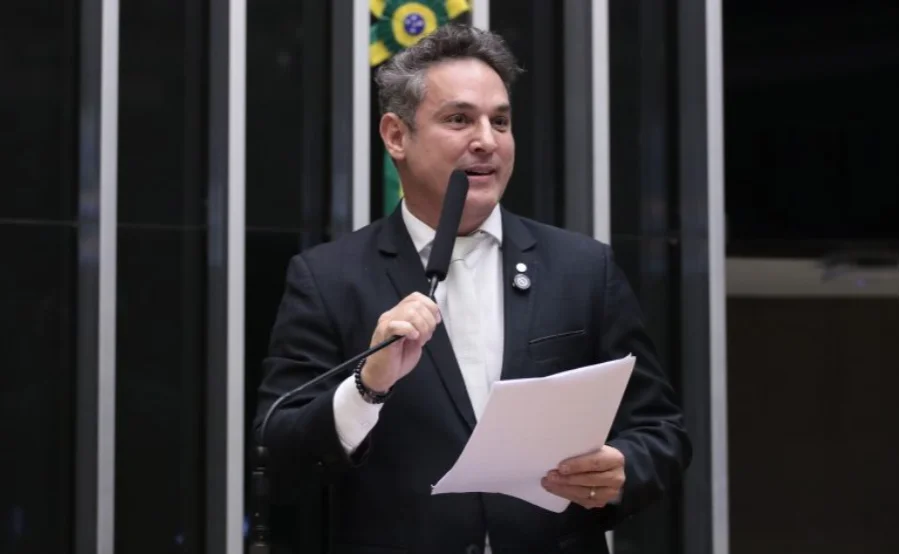 Deputado Zucco discursa na Câmara dos Deputados, em Brasília, durante sessão de comissão. Ele segura papéis e um microfone, com o brasão do Brasil ao fundo. Imagem relacionada à aprovação do projeto de lei de indenização de armas.