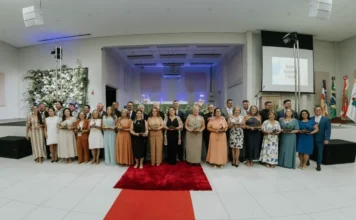 Câmara de Araranguá entrega Troféu Alzira Rabelo Elias Fotografia em plano geral das 15 mulheres homenageadas com o 24º Troféu Alzira Rabelo Elias, posando lado a lado com vereadores e autoridades de Araranguá. O grupo está organizado em frente a um palco no Centro Multiuso, posicionado sobre um tapete vermelho central. As homenageadas seguram seus troféus, e ao fundo, à direita, um telão exibe a logomarca do evento.