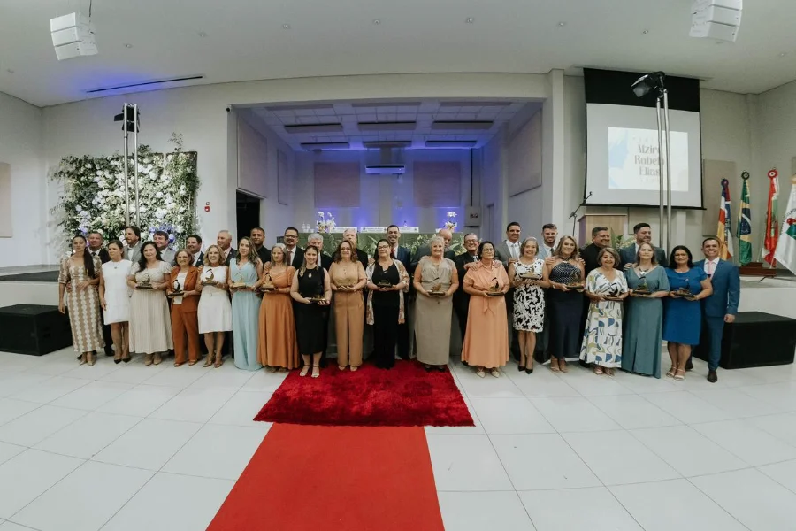 Fotografia em plano geral das 15 mulheres homenageadas com o 24º Troféu Alzira Rabelo Elias, posando lado a lado com vereadores e autoridades de Araranguá. O grupo está organizado em frente a um palco no Centro Multiuso, posicionado sobre um tapete vermelho central. As homenageadas seguram seus troféus, e ao fundo, à direita, um telão exibe a logomarca do evento.