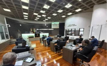 Câmara de Araranguá vota melhorias na BR-101 e segurança Ampla visão do plenário da Câmara de Vereadores de Araranguá durante sessão ordinária. Parlamentares vestindo trajes formais estão sentados em bancadas individuais de madeira, equipadas com monitores e documentos. Ao fundo, a mesa diretora conduz os trabalhos sob um letreiro com o nome da instituição, ladeado pelas bandeiras oficiais. O ambiente possui piso de madeira, teto quadriculado com iluminação embutida e uma inscrição bíblica na parede lateral.