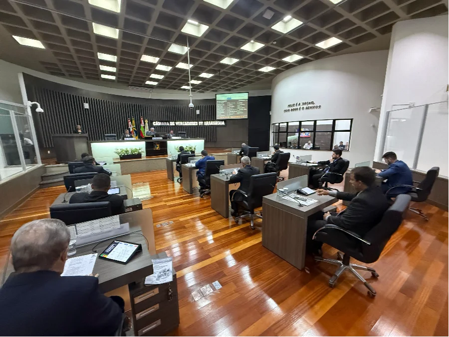 Ampla visão do plenário da Câmara de Vereadores de Araranguá durante sessão ordinária. Parlamentares vestindo trajes formais estão sentados em bancadas individuais de madeira, equipadas com monitores e documentos. Ao fundo, a mesa diretora conduz os trabalhos sob um letreiro com o nome da instituição, ladeado pelas bandeiras oficiais. O ambiente possui piso de madeira, teto quadriculado com iluminação embutida e uma inscrição bíblica na parede lateral.