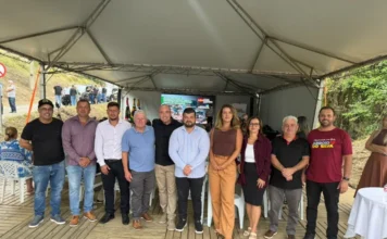 Câmara de Arroio do Silva destaca início de obra na SC-447 Vereadores e autoridades de Balneário Arroio do Silva reunidos sob uma tenda durante a solenidade de assinatura da ordem de serviço para a construção da ciclovia na rodovia SC-447. Ao fundo, tela exibe informações sobre investimentos em infraestrutura e mobilidade urbana.