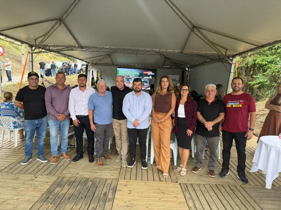 Vereadores e autoridades de Balneário Arroio do Silva reunidos sob uma tenda durante a solenidade de assinatura da ordem de serviço para a construção da ciclovia na rodovia SC-447. Ao fundo, tela exibe informações sobre investimentos em infraestrutura e mobilidade urbana.