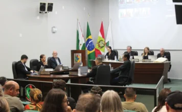 Câmara de Forquilhinha debate IPTU e aprova novos projetos Uma fotografia tirada da plateia para o palco de uma sessão de câmara municipal em andamento. No palco, uma mesa de conselho está ocupada por cerca de dez pessoas em trajes formais, cada uma com um notebook e placas de identificação. Há quatro bandeiras oficiais penduradas atrás deles (incluindo as bandeiras do Brasil, de Santa Catarina e duas municipais). Uma grande tela de projeção atrás da mesa exibe participantes remotos em grade e texto com uma contagem de tempo. A plateia está sentada em cadeiras em primeiro plano, observando a sessão.