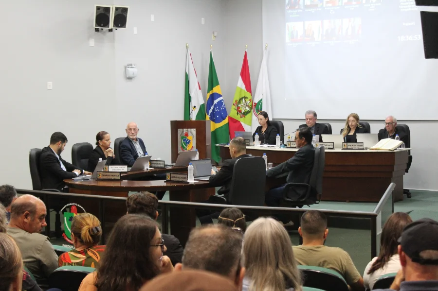 Uma fotografia tirada da plateia para o palco de uma sessão de câmara municipal em andamento. No palco, uma mesa de conselho está ocupada por cerca de dez pessoas em trajes formais, cada uma com um notebook e placas de identificação. Há quatro bandeiras oficiais penduradas atrás deles (incluindo as bandeiras do Brasil, de Santa Catarina e duas municipais). Uma grande tela de projeção atrás da mesa exibe participantes remotos em grade e texto com uma contagem de tempo. A plateia está sentada em cadeiras em primeiro plano, observando a sessão.