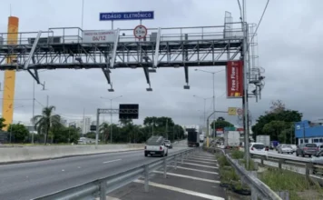 Câmara debate falhas e multas no pedágio free-flow Pórtico metálico de sistema de pedágio free-flow instalado sobre rodovia com sensores de cobrança eletrônica e placa indicativa de livre passagem em dia nublado.