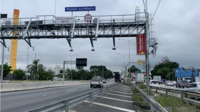 Câmara debate falhas e multas no pedágio free-flow Pórtico metálico de sistema de pedágio free-flow instalado sobre rodovia com sensores de cobrança eletrônica e placa indicativa de livre passagem em dia nublado.
