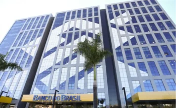 Câmara debate mudanças nas carreiras do Banco do Brasil Vista em ângulo baixo da fachada moderna de um edifício do Banco do Brasil, com grandes janelas de vidro que refletem o céu. Na base do prédio, logo acima da entrada, destaca-se o letreiro azul e amarelo com o nome da instituição. À frente, encontram-se palmeiras e vasos de plantas, compondo a sede da estatal que é o centro do debate sobre a reestruturação de carreiras e condições laborais no Parlamento.