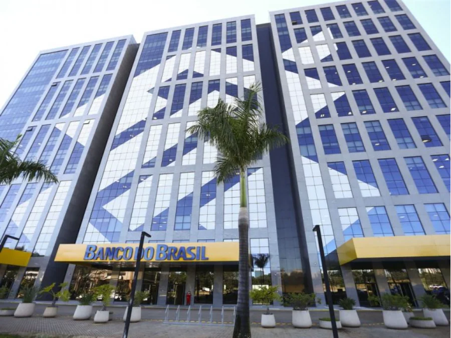 Vista em ângulo baixo da fachada moderna de um edifício do Banco do Brasil, com grandes janelas de vidro que refletem o céu. Na base do prédio, logo acima da entrada, destaca-se o letreiro azul e amarelo com o nome da instituição. À frente, encontram-se palmeiras e vasos de plantas, compondo a sede da estatal que é o centro do debate sobre a reestruturação de carreiras e condições laborais no Parlamento.