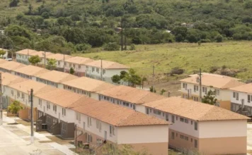 Câmara debate penas por atrasos no Minha Casa, Minha Vida Vista aérea de um grande e organizado complexo habitacional recém-construído, composto por múltiplas filas longas e paralelas de casas geminadas idênticas de dois andares. As moradias possuem telhados de telhas de barro laranja e fachadas pintadas em tons suaves de bege, creme e verde-pálido, com áreas pavimentadas e postes de luz. O local está situado em uma área de transição, com um vasto campo gramado em primeiro plano e, ao fundo, uma encosta densamente coberta por vegetação verde e árvores.