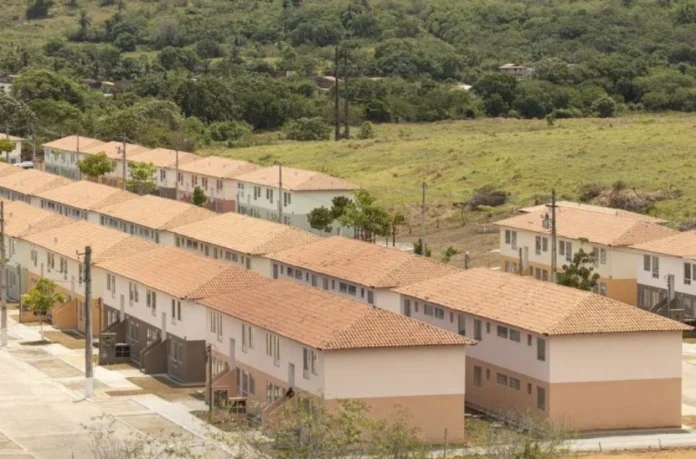 Câmara debate penas por atrasos no Minha Casa, Minha Vida Vista aérea de um grande e organizado complexo habitacional recém-construído, composto por múltiplas filas longas e paralelas de casas geminadas idênticas de dois andares. As moradias possuem telhados de telhas de barro laranja e fachadas pintadas em tons suaves de bege, creme e verde-pálido, com áreas pavimentadas e postes de luz. O local está situado em uma área de transição, com um vasto campo gramado em primeiro plano e, ao fundo, uma encosta densamente coberta por vegetação verde e árvores.