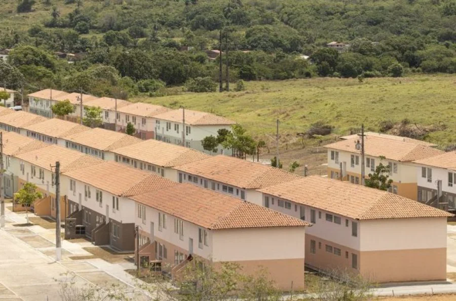Vista aérea de um grande e organizado complexo habitacional recém-construído, composto por múltiplas filas longas e paralelas de casas geminadas idênticas de dois andares. As moradias possuem telhados de telhas de barro laranja e fachadas pintadas em tons suaves de bege, creme e verde-pálido, com áreas pavimentadas e postes de luz. O local está situado em uma área de transição, com um vasto campo gramado em primeiro plano e, ao fundo, uma encosta densamente coberta por vegetação verde e árvores.