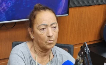 Campanha arrecada fundos para cadeira de rodas elétrica Dona Cecília, uma senhora idosa de expressão séria e olhar atento, durante sua entrevista nos estúdios da Rádio Amorim FM. Ela está posicionada de perfil, em frente a um microfone com a logomarca da emissora, vestindo uma blusa cinza. Ao fundo, é possível observar parte da estrutura acústica do estúdio e um monitor de televisão.