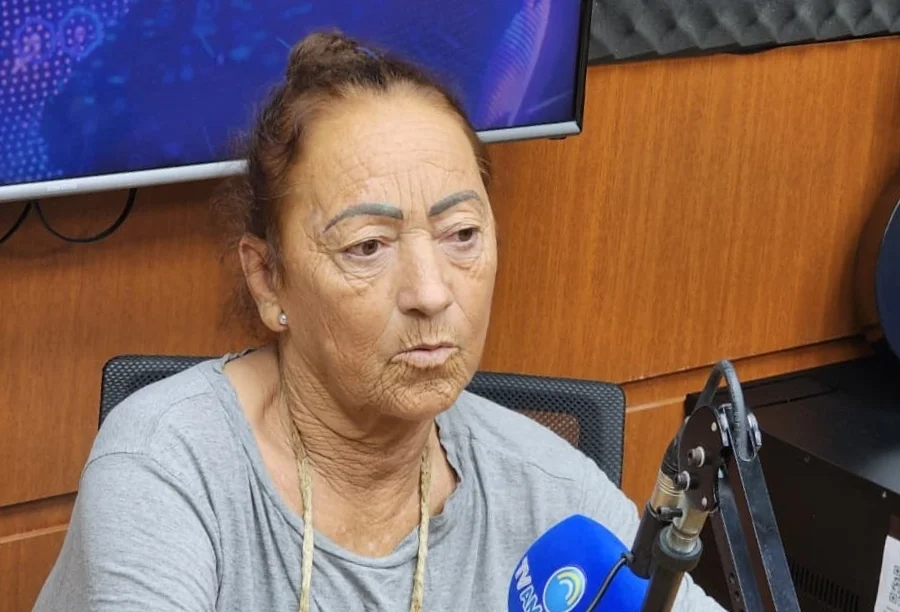 _Campanha arrecada fundos para cadeira de rodas elétrica Dona Cecília, uma senhora idosa de expressão séria e olhar atento, durante sua entrevista nos estúdios da Rádio Amorim FM. Ela está posicionada de perfil, em frente a um microfone com a logomarca da emissora, vestindo uma blusa cinza. Ao fundo, é possível observar parte da estrutura acústica do estúdio e um monitor de televisão.