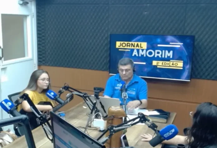 Cantora Thalyta Silva fala sobre chamado e fé na rádio Cantora gospel Thalyta Silva segura um violão durante entrevista nos estúdios da Rádio 102.9 Amorim FM. Ao fundo, monitor exibe a identidade visual do Jornal Amorim 2ª Edição em um ambiente de estúdio profissional.