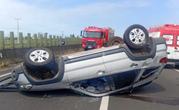 Capotamento na BR-101 deixa quatro feridos em Sombrio Fotografia em plano médio mostra um veículo Fiat Palio Weekend de cor prata capotado com as rodas para cima, ocupando uma das faixas da rodovia BR-101. O carro apresenta danos estruturais visíveis no teto e nas janelas laterais. Ao fundo, em solo asfáltico, veem-se um caminhão vermelho do Corpo de Bombeiros (ABTR) e uma ambulância do SAMU estacionados para o atendimento da ocorrência. O cenário é de dia claro, com barreiras de concreto e uma grade de proteção verde ao lado da pista.