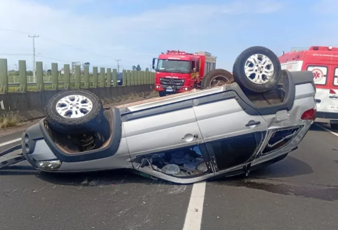 Capotamento na BR-101 deixa quatro feridos em Sombrio Fotografia em plano médio mostra um veículo Fiat Palio Weekend de cor prata capotado com as rodas para cima, ocupando uma das faixas da rodovia BR-101. O carro apresenta danos estruturais visíveis no teto e nas janelas laterais. Ao fundo, em solo asfáltico, veem-se um caminhão vermelho do Corpo de Bombeiros (ABTR) e uma ambulância do SAMU estacionados para o atendimento da ocorrência. O cenário é de dia claro, com barreiras de concreto e uma grade de proteção verde ao lado da pista.