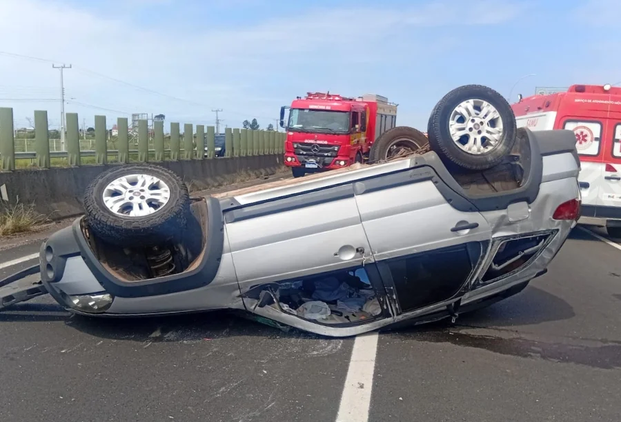 Fotografia em plano médio mostra um veículo Fiat Palio Weekend de cor prata capotado com as rodas para cima, ocupando uma das faixas da rodovia BR-101. O carro apresenta danos estruturais visíveis no teto e nas janelas laterais. Ao fundo, em solo asfáltico, veem-se um caminhão vermelho do Corpo de Bombeiros (ABTR) e uma ambulância do SAMU estacionados para o atendimento da ocorrência. O cenário é de dia claro, com barreiras de concreto e uma grade de proteção verde ao lado da pista.