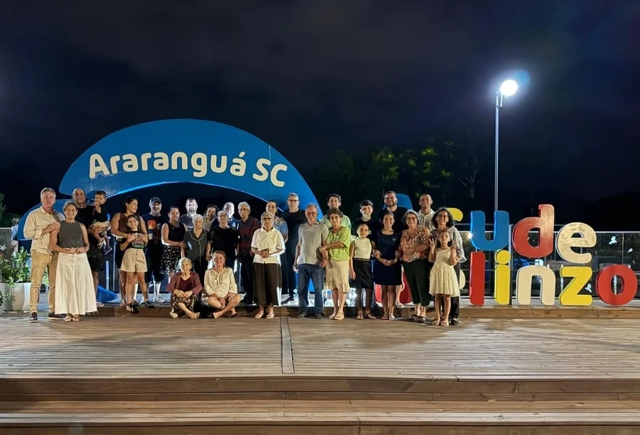Uma grande foto de família reúne Domingos e Salete Barbosa com filhos, netos e bisnetos no deck de madeira à noite. Ao fundo, o letreiro azul com a inscrição Araranguá SC e luzes do Parque Belinzoni.
