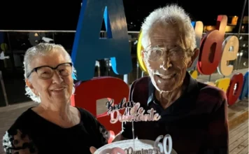 Casal celebra 60 anos de união no Parque Belinzoni Domingos e Salete Barbosa sorriem para a foto celebrando 60 anos de casados. Eles seguram um bolo personalizado de Bodas de Diamante com letreiros coloridos ao fundo no Parque Belinzoni em Araranguá, SC.