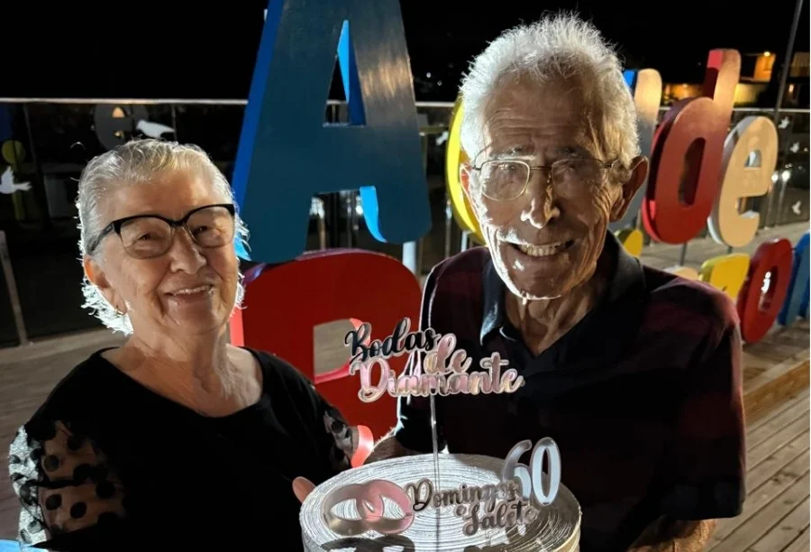Domingos e Salete Barbosa sorriem para a foto celebrando 60 anos de casados. Eles seguram um bolo personalizado de Bodas de Diamante com letreiros coloridos ao fundo no Parque Belinzoni em Araranguá, SC.