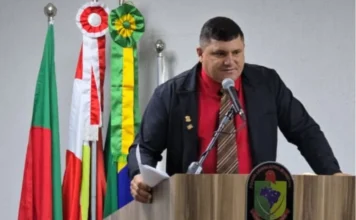 Chico do Maracujá assume a presidência da Câmara de Sombrio Marilon Cardoso, o Chico do Maracujá, no púlpito da Câmara de Vereadores de Sombrio após eleição como presidente. O vereador veste camisa vermelha e jaqueta preta, com bandeiras oficiais e o brasão municipal ao fundo, falando ao microfone.