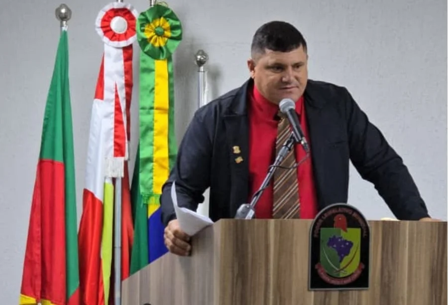 Chico do Maracujá assume a presidência da Câmara de Sombrio Marilon Cardoso, o Chico do Maracujá, no púlpito da Câmara de Vereadores de Sombrio após eleição como presidente. O vereador veste camisa vermelha e jaqueta preta, com bandeiras oficiais e o brasão municipal ao fundo, falando ao microfone.