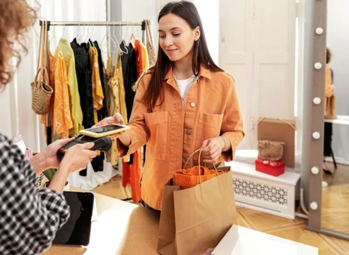 Comércio de Araranguá amplia horário para o Dia da Mulher Mulher jovem de cabelos escuros e casaco alaranjado sorri ao realizar um pagamento por aproximação com o celular em uma loja de roupas. Ela segura uma sacola de papel pardo enquanto a vendedora segura a máquina de cartões. Ao fundo, um arara com peças de vestuário coloridas e um espelho de camarim completam o ambiente de compras no comércio de Araranguá.