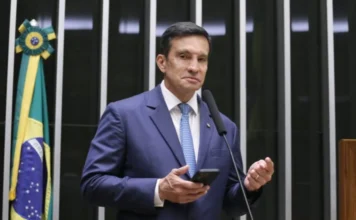 Comissão aprova rastreamento digital contra garimpo ilegal O deputado federal Sidney Leite (PSD-AM) fala no púlpito da Câmara dos Deputados sobre o projeto de rastreabilidade digital do ouro. Ele segura um smartphone e está de terno azul. No fundo, painéis de votação e bandeira do Brasil.