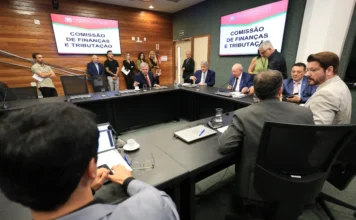 Comissão aprova redução de ICMS para setor agropecuário Fotografia em ângulo médio de uma reunião presencial da Comissão de Finanças e Tributação. Vários homens de terno e algumas mulheres estão sentados ao redor de uma grande mesa em forma de U. Duas telas de TV nas paredes exibem o título da comissão. Laptops, copos de água e microfones estão sobre a mesa. A foto é tirada de trás de duas pessoas em primeiro plano.