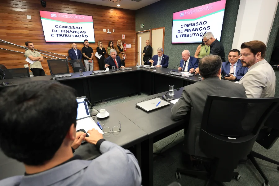 Fotografia em ângulo médio de uma reunião presencial da Comissão de Finanças e Tributação. Vários homens de terno e algumas mulheres estão sentados ao redor de uma grande mesa em forma de U. Duas telas de TV nas paredes exibem o título da comissão. Laptops, copos de água e microfones estão sobre a mesa. A foto é tirada de trás de duas pessoas em primeiro plano.