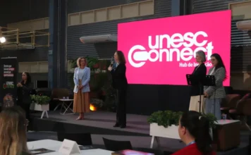 Conectando Mulheres reúne 400 participantes em Criciúma Palco do evento Conectando Mulheres do Extremo Sul no Centro de Inovação Criciúma (CRIO). A pastora Viviane Martinello, vestindo um blazer quadriculado, fala ao microfone em frente a um grande telão LED rosa com o logotipo 'unesc Connect Hub de Inovação'. Ao seu lado, com a pose de escuta, está a reitora em exercício da Unesc, Gisele Silveira Coelho Lopes (em pé à esquerda), e a coordenadora do Hub, Jaqueline Bitencourt Lopes (em pé à direita). Uma intérprete de LIBRAS gesticula à esquerda. Parte da plateia assiste.