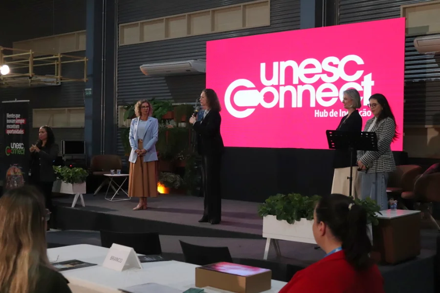 Palco do evento Conectando Mulheres do Extremo Sul no Centro de Inovação Criciúma (CRIO). A pastora Viviane Martinello, vestindo um blazer quadriculado, fala ao microfone em frente a um grande telão LED rosa com o logotipo 'unesc Connect Hub de Inovação'. Ao seu lado, com a pose de escuta, está a reitora em exercício da Unesc, Gisele Silveira Coelho Lopes (em pé à esquerda), e a coordenadora do Hub, Jaqueline Bitencourt Lopes (em pé à direita). Uma intérprete de LIBRAS gesticula à esquerda. Parte da plateia assiste.