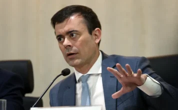 Conflito no Irã pode frear queda de juros no Brasil Fotografia em plano médio do secretário do Tesouro Nacional, Rogério Ceron, em uma mesa de conferência. Ele é um homem de pele clara, cabelos escuros e curtos, veste um terno cinza escuro sobre uma camisa branca e usa uma gravata azul-clara. Ceron está posicionado à frente de um microfone e gesticula com a mão direita aberta, demonstrando ênfase em sua fala sobre o cenário econômico e o conflito no Irã. À sua frente, uma placa de identificação exibe seu nome em letras pretas: "ROGÉRIO CERON".