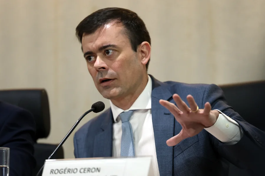 Fotografia em plano médio do secretário do Tesouro Nacional, Rogério Ceron, em uma mesa de conferência. Ele é um homem de pele clara, cabelos escuros e curtos, veste um terno cinza escuro sobre uma camisa branca e usa uma gravata azul-clara. Ceron está posicionado à frente de um microfone e gesticula com a mão direita aberta, demonstrando ênfase em sua fala sobre o cenário econômico e o conflito no Irã. À sua frente, uma placa de identificação exibe seu nome em letras pretas: 