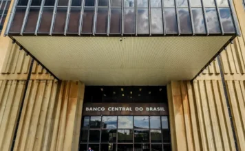 Copom decide hoje primeiro corte da Selic em dois anos Fotografia de ângulo baixo da fachada e entrada principal do edifício-sede do Banco Central do Brasil em Brasília. A imagem destaca a placa de metal com o nome "BANCO CENTRAL DO BRASIL", situada acima das portas de vidro e sob uma grande marquise de metal ripado, com a estrutura de concreto brutalista visível ao redor.