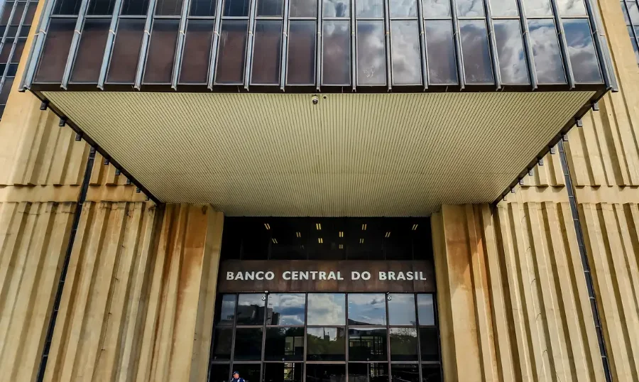 Fotografia de ângulo baixo da fachada e entrada principal do edifício-sede do Banco Central do Brasil em Brasília. A imagem destaca a placa de metal com o nome 