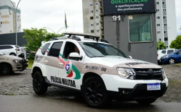 Crianças são resgatadas em local de tráfico em Criciúma Viatura Renault Duster da Polícia Militar de Santa Catarina (PMSC) estacionada em frente a um prédio de qualificação profissional em dia nublado. A foto, em close-up, mostra o fardamento do policial dentro do veículo, o logotipo da PMSC, a identificação de emergência 190 e a placa QJS4C94. Imagem ilustrativa para notícias sobre ações policiais, segurança pública e combate ao tráfico de drogas em SC, incluindo Criciúma.