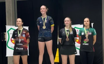 Criciúma conquista 16 medalhas no tênis de mesa estadual Quatro jovens atletas do sexo feminino de pé em um pódio de premiação em um ambiente interno. Elas estão organizadas da esquerda para a direita em níveis de segundo, primeiro e dois de terceiro lugar. Cada atleta usa uma medalha no pescoço e sorri para a câmera. Elas vestem uniformes de equipe diferentes e coloridos com logos de patrocinadores. Atrás delas, uma cortina preta e banners da federação esportiva são visíveis.