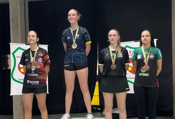 Criciúma conquista 16 medalhas no tênis de mesa estadual Quatro jovens atletas do sexo feminino de pé em um pódio de premiação em um ambiente interno. Elas estão organizadas da esquerda para a direita em níveis de segundo, primeiro e dois de terceiro lugar. Cada atleta usa uma medalha no pescoço e sorri para a câmera. Elas vestem uniformes de equipe diferentes e coloridos com logos de patrocinadores. Atrás delas, uma cortina preta e banners da federação esportiva são visíveis.