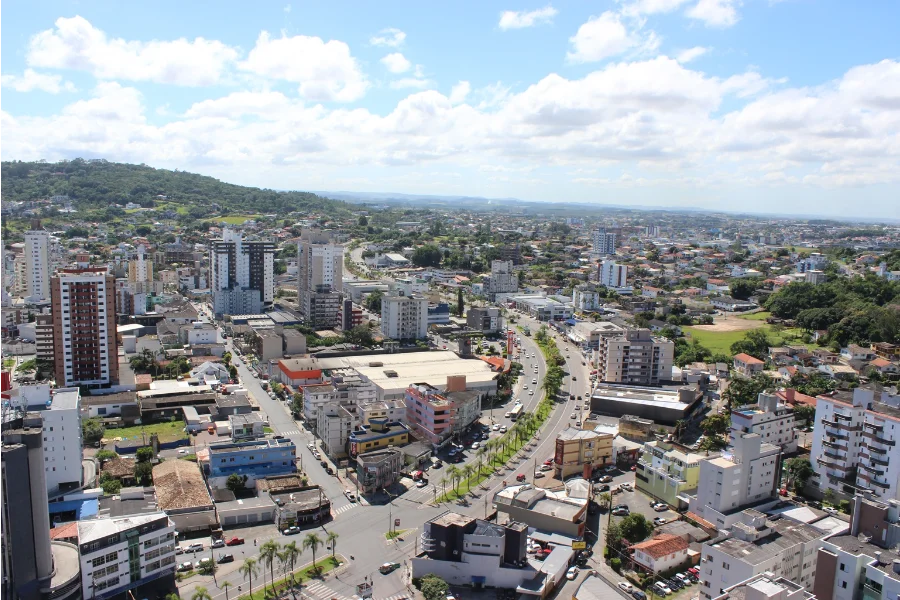 Vista aérea panorâmica da área urbana de Criciúma, em Santa Catarina, exibindo densidade de edifícios residenciais e comerciais, avenidas movimentadas com veículos e vegetação ao fundo sob um céu claro com nuvens. A imagem ilustra o desenvolvimento e a infraestrutura do município mencionado no Ranking de Competitividade.