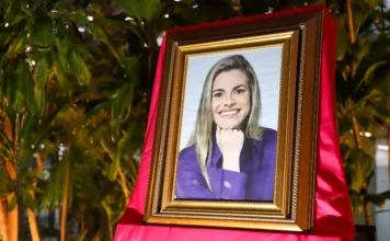 Criciúma homenageia 33 mulheres com Medalha Thati Teixeira Um retrato de uma mulher sorridente com longos cabelos loiros e uma blusa roxa, com a mão apoiada sob o queixo, é apresentado em uma moldura dourada ornamentada. A moldura está apoiada sobre um suporte coberto por um tecido vermelho drapeado. O fundo é um ambiente interno com folhagem verde exuberante e iluminação quente.