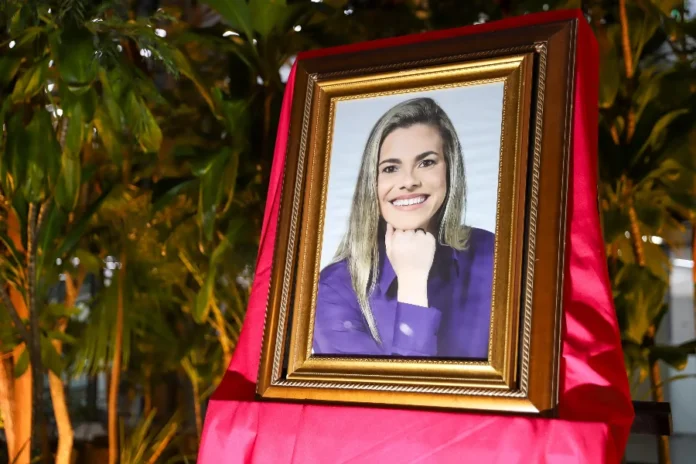 Criciúma homenageia 33 mulheres com Medalha Thati Teixeira Um retrato de uma mulher sorridente com longos cabelos loiros e uma blusa roxa, com a mão apoiada sob o queixo, é apresentado em uma moldura dourada ornamentada. A moldura está apoiada sobre um suporte coberto por um tecido vermelho drapeado. O fundo é um ambiente interno com folhagem verde exuberante e iluminação quente.