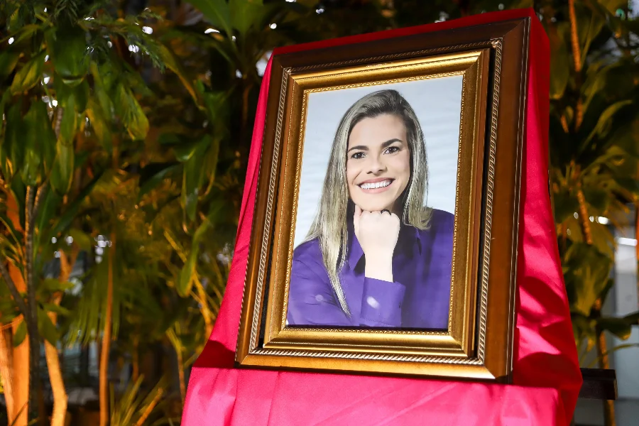 Um retrato de uma mulher sorridente com longos cabelos loiros e uma blusa roxa, com a mão apoiada sob o queixo, é apresentado em uma moldura dourada ornamentada. A moldura está apoiada sobre um suporte coberto por um tecido vermelho drapeado. O fundo é um ambiente interno com folhagem verde exuberante e iluminação quente.