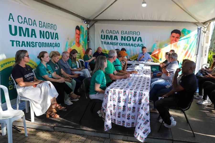 Encontro de escuta ativa do Gabinete Itinerante da Prefeitura de Criciúma na região da Cidade Mineira. Prefeito Vagner Espindola, microfone na mão, lidera diálogo com moradores sob tenda. Banners com a frase 'A Cada Bairro Uma Nova Transformação' são visíveis.