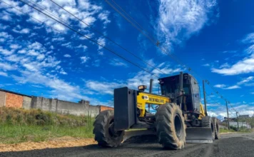 Criciúma inicia pavimentação de R$ 1,2 milhão no Santo Antônio Motoniveladora amarela New Holland operando em obra de pavimentação asfáltica em Criciúma. A máquina pesada está nivelando uma camada de asfalto fresco em uma estrada secundária, com um muro de tijolos e postes de luz ao fundo sob um céu azul vibrante com nuvens.