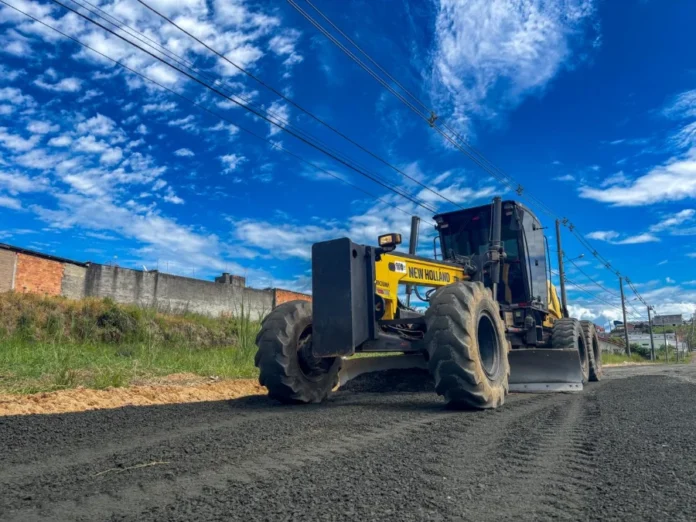 Criciúma inicia pavimentação de R$ 1,2 milhão no Santo Antônio Motoniveladora amarela New Holland operando em obra de pavimentação asfáltica em Criciúma. A máquina pesada está nivelando uma camada de asfalto fresco em uma estrada secundária, com um muro de tijolos e postes de luz ao fundo sob um céu azul vibrante com nuvens.