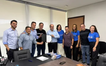 Criciúma investe R$ 650 mil em estrutura para autistas Fotografia em plano médio capturada no Paço Municipal Marcos Rovaris. Ao centro da imagem, o prefeito de Criciúma, Vagner Espindola, e o presidente da AMA, José Augusto Freitas, seguram conjuntamente uma pasta preta com o documento oficial do repasse de R$ 650 mil. Eles estão acompanhados por outros oito representantes do governo e da instituição, dispostos em pé atrás de uma mesa de reunião. Alguns integrantes da associação vestem camisetas azuis com a logomarca da AMA. O ambiente é um escritório formal com paredes brancas, janelas com persianas e equipamentos de escritório sobre a mesa.