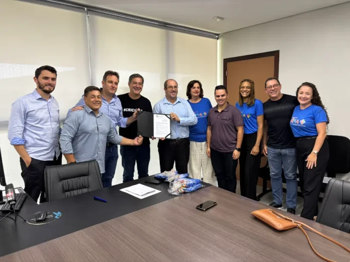 Criciúma investe R$ 650 mil em estrutura para autistas Fotografia em plano médio capturada no Paço Municipal Marcos Rovaris. Ao centro da imagem, o prefeito de Criciúma, Vagner Espindola, e o presidente da AMA, José Augusto Freitas, seguram conjuntamente uma pasta preta com o documento oficial do repasse de R$ 650 mil. Eles estão acompanhados por outros oito representantes do governo e da instituição, dispostos em pé atrás de uma mesa de reunião. Alguns integrantes da associação vestem camisetas azuis com a logomarca da AMA. O ambiente é um escritório formal com paredes brancas, janelas com persianas e equipamentos de escritório sobre a mesa.