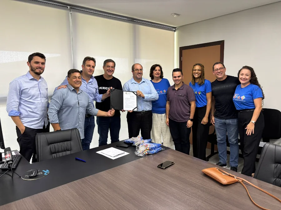 Criciúma investe R$ 650 mil em estrutura para autistas Fotografia em plano médio capturada no Paço Municipal Marcos Rovaris. Ao centro da imagem, o prefeito de Criciúma, Vagner Espindola, e o presidente da AMA, José Augusto Freitas, seguram conjuntamente uma pasta preta com o documento oficial do repasse de R$ 650 mil. Eles estão acompanhados por outros oito representantes do governo e da instituição, dispostos em pé atrás de uma mesa de reunião. Alguns integrantes da associação vestem camisetas azuis com a logomarca da AMA. O ambiente é um escritório formal com paredes brancas, janelas com persianas e equipamentos de escritório sobre a mesa.